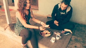 Dues persones assegudes en un graó de formigó davant d'un edifici amb una paret vermella i blanca, compartint un àpat amb pa, un plat i gots.