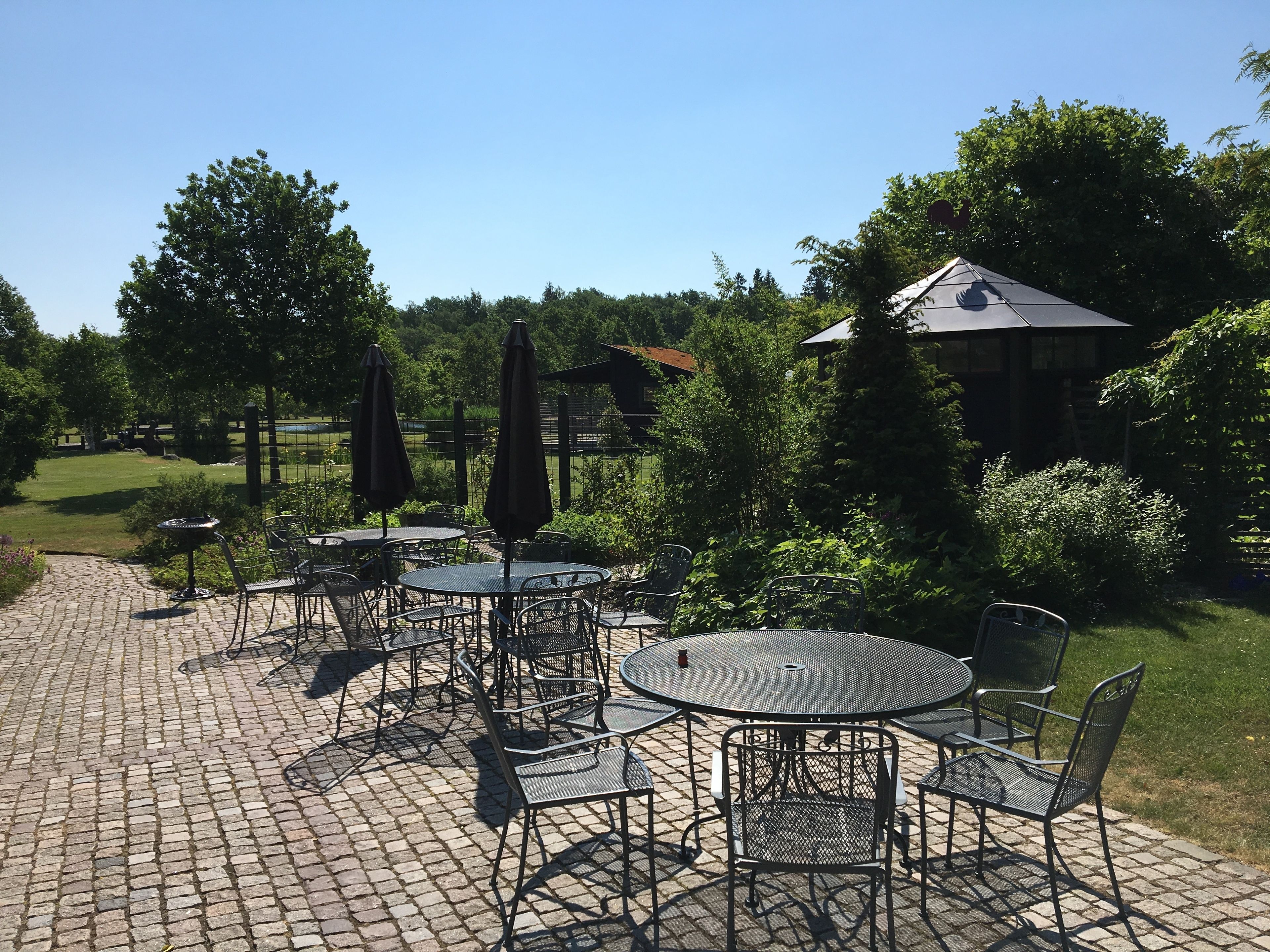 Ein Patio mit Sitzgelegenheiten im Freien, darunter runde Tische und Stühle, umgeben von Grünpflanzen und einem Backsteinweg.