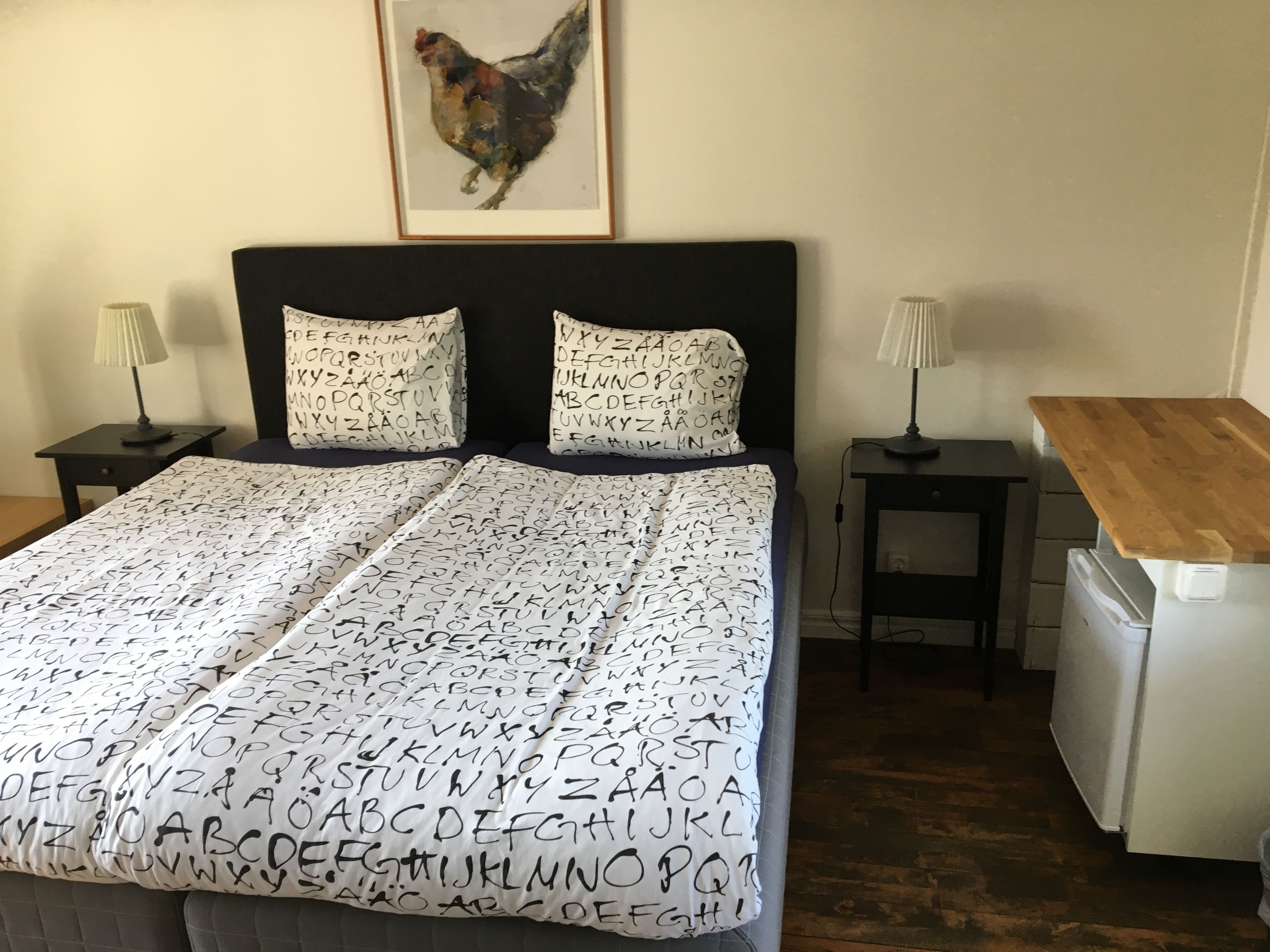 A bedroom with a bed, pillows with alphabet patterns, a framed artwork of a chicken, bedside tables with lamps, and a wooden desk with a chair.