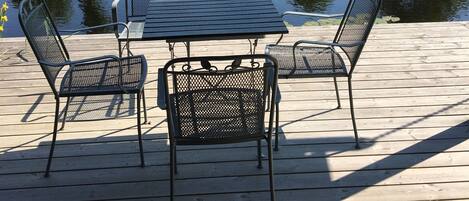 A wooden deck with a table and chairs overlooking a calm pond with a wooden dock and trees in the background.