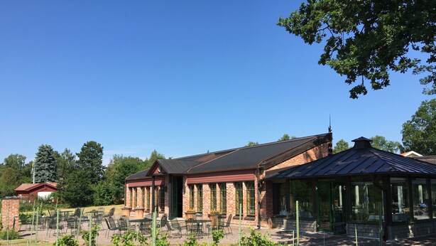 Un bâtiment en briques doté d’une véranda en verre, entouré d’un jardin où sont alignées de jeunes plantes.
