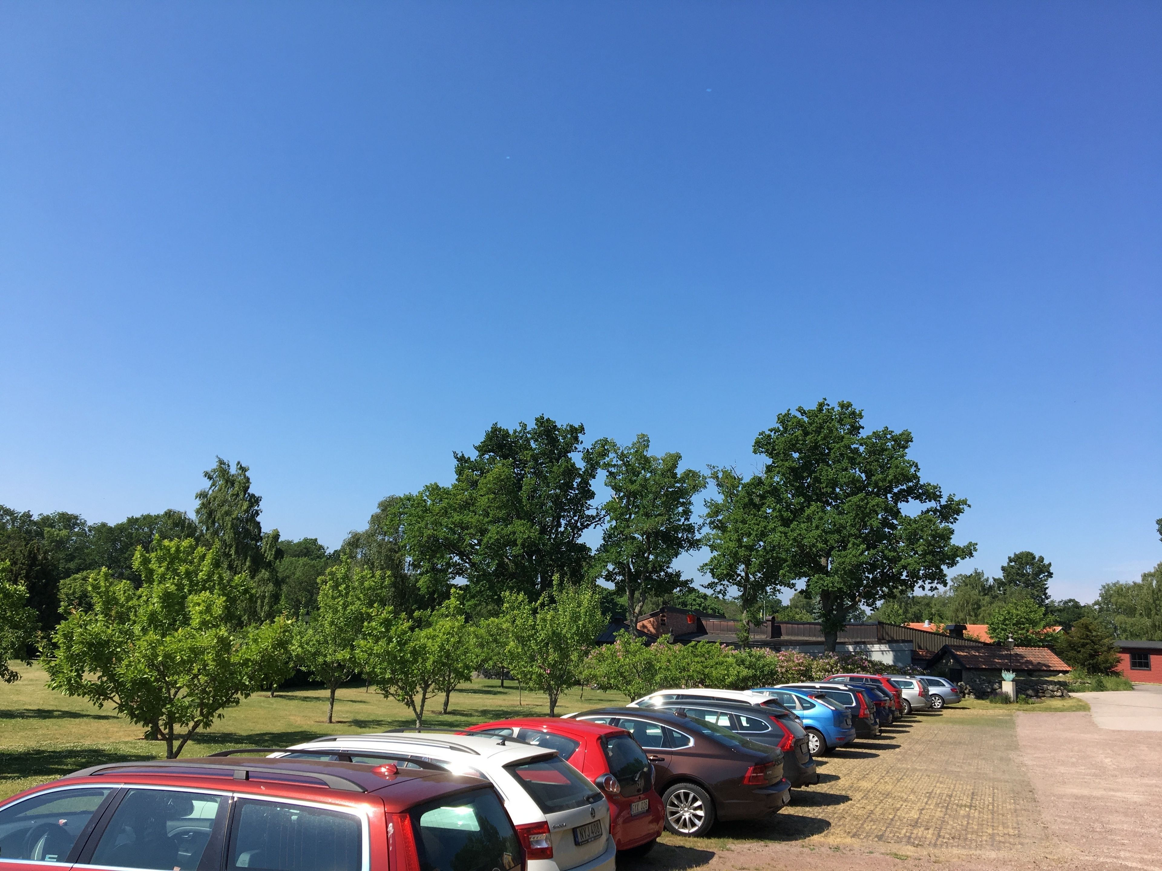 A parking lot with multiple cars parked in rows, surrounded by trees and a building with a red roof.