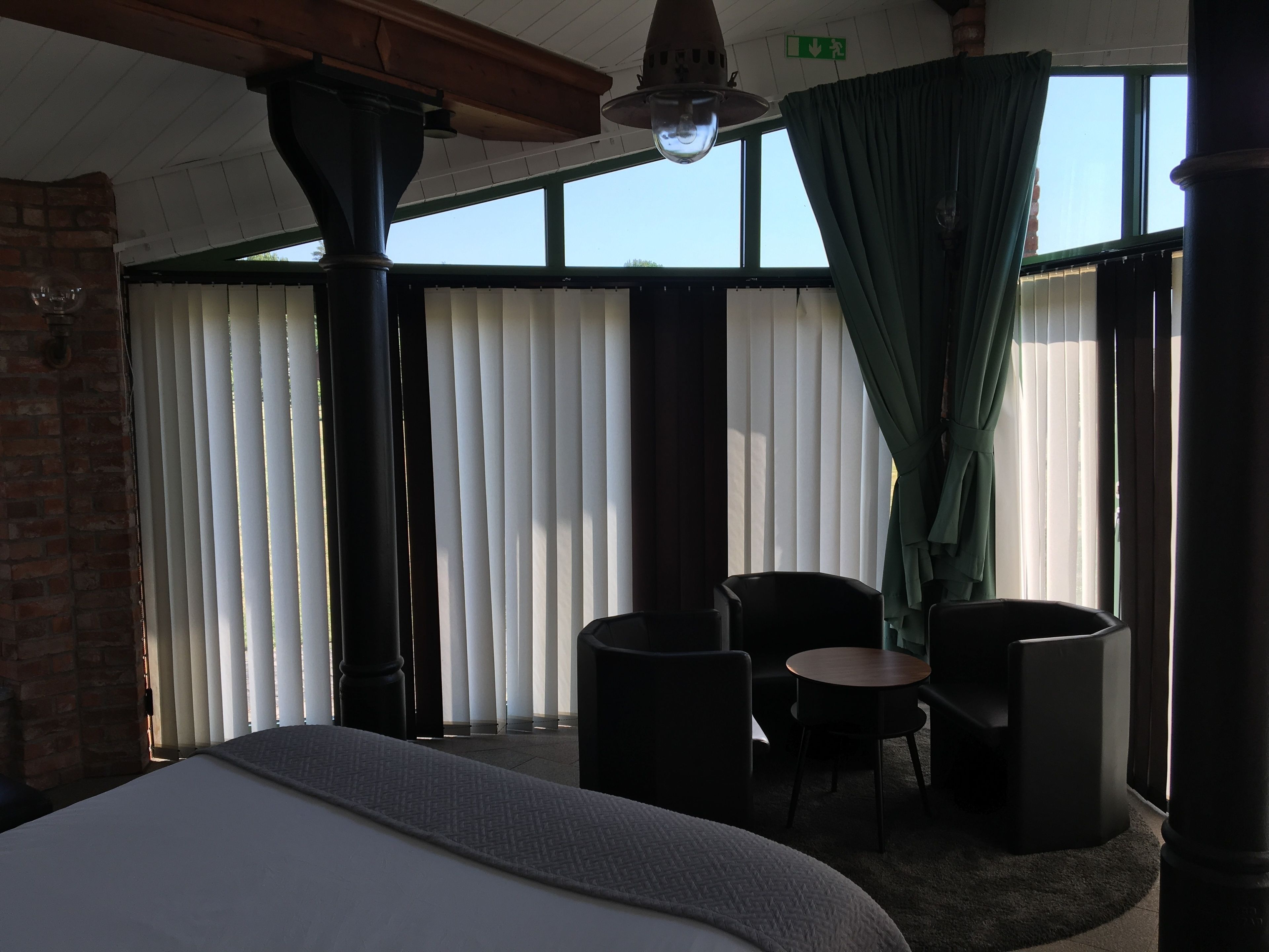 A hotel room with a large bed, two chairs, a small table, and a view of the outdoors through large windows with curtains.