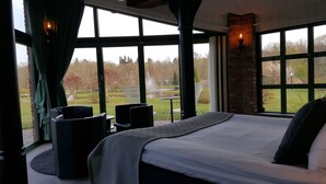Une chambre avec un grand lit, deux fauteuils, une table et une vue sur un parc avec un étang et des arbres.