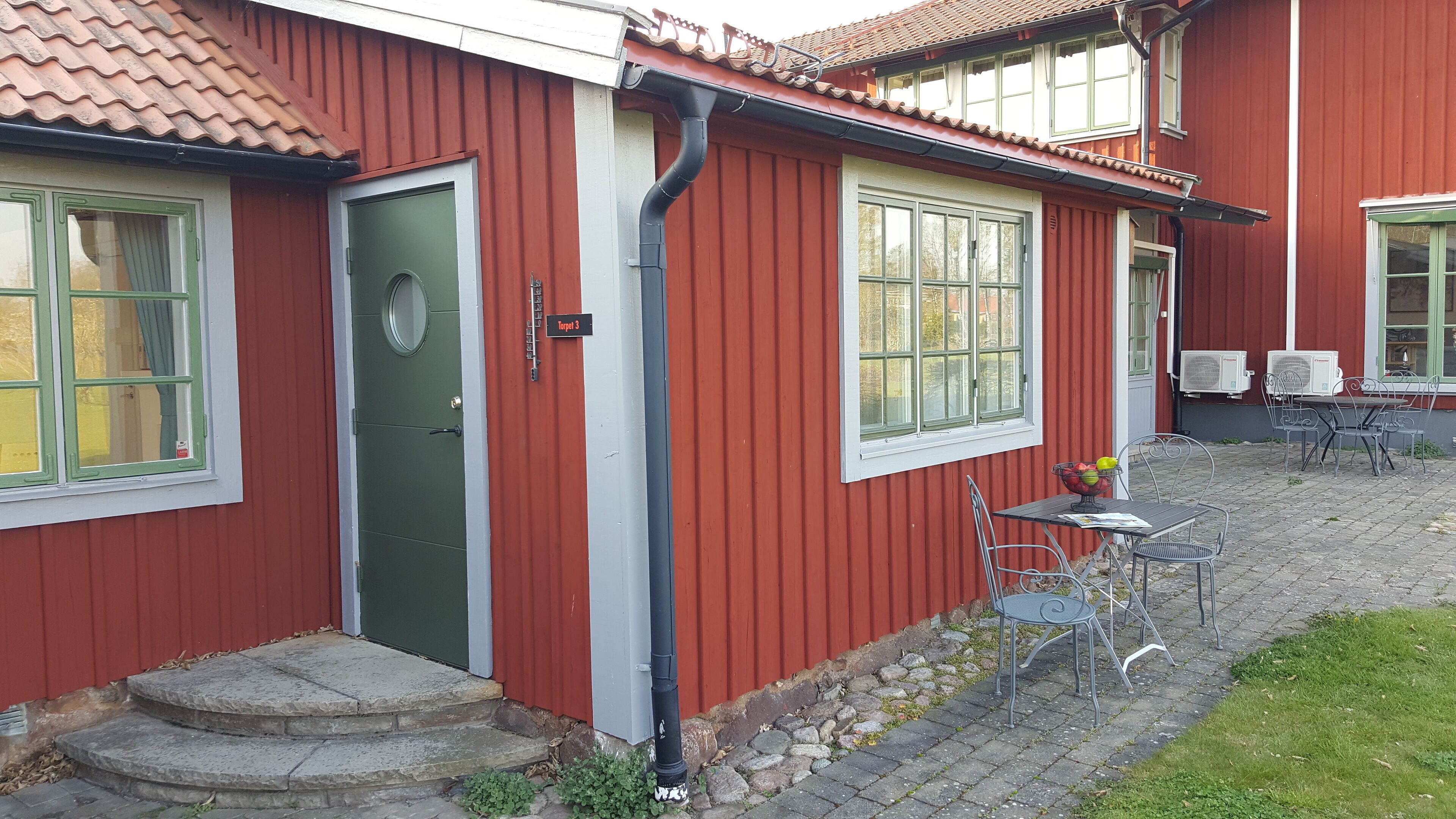 A red building with a green door, a small outdoor seating area with metal chairs and a table, and two air conditioning units on the side.