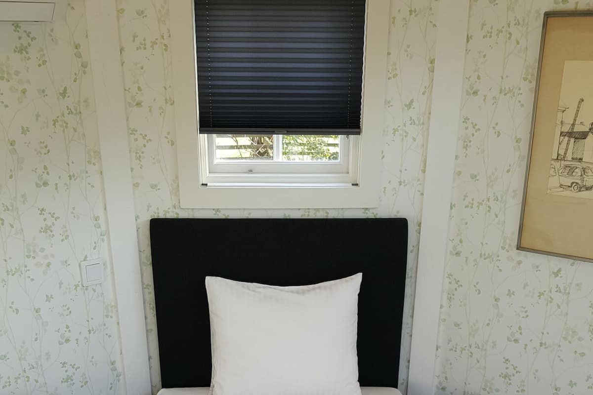 Una cama individual con cabecera negra, ropa de cama blanca y dos toallas dobladas al pie. Una ventana con persianas está sobre la cama, y un cuadro enmarcado cuelga en la pared a la derecha.