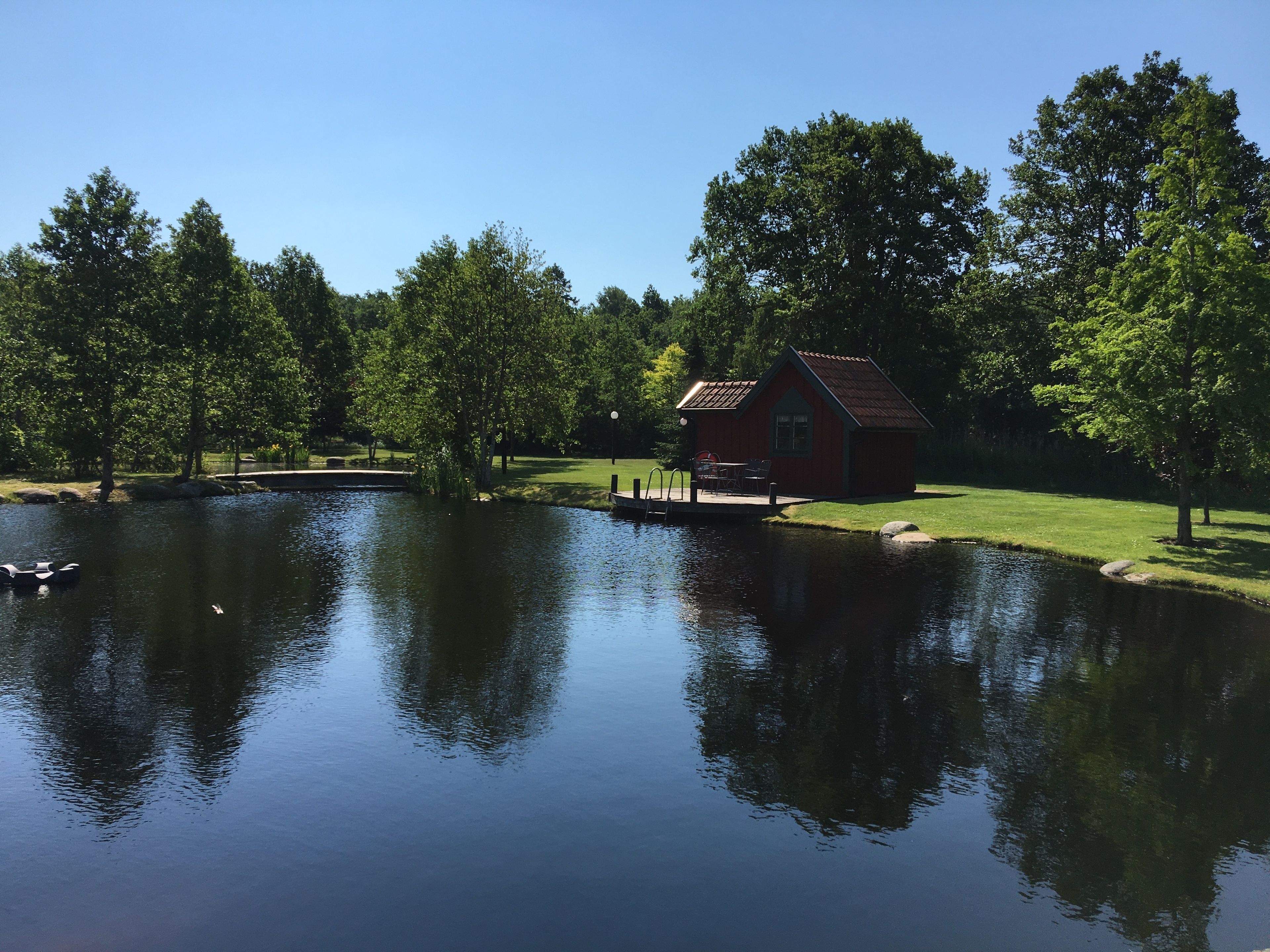 A tranquil lakeside setting with a small red cabin, surrounded by lush greenery and a clear blue sky.