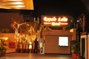 L’entrée d’un hôtel, avec une enseigne au néon indiquant « Royale Lait Hotel ». Des plantes en pot et une personne se tiennent près de l’entrée.