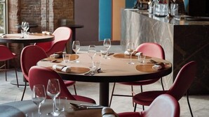 Interior de restaurante con mesas redondas, sillas rojas y una zona de bar con estantes llenos de botellas.