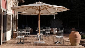 Un patio exterior con mesas y sillas, un gran paraguas y un edificio rústico al fondo.