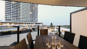 Un balcone con sedie in vimini e un tavolo apparecchiato con bicchieri, con vista su un'area parcheggio e un edificio alto.