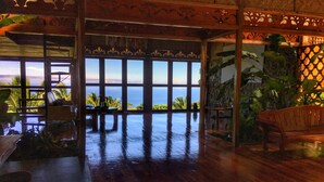 Une pièce spacieuse dotée de grandes fenêtres donnant sur l’océan, d’un plancher en bois et de plantes tropicales.