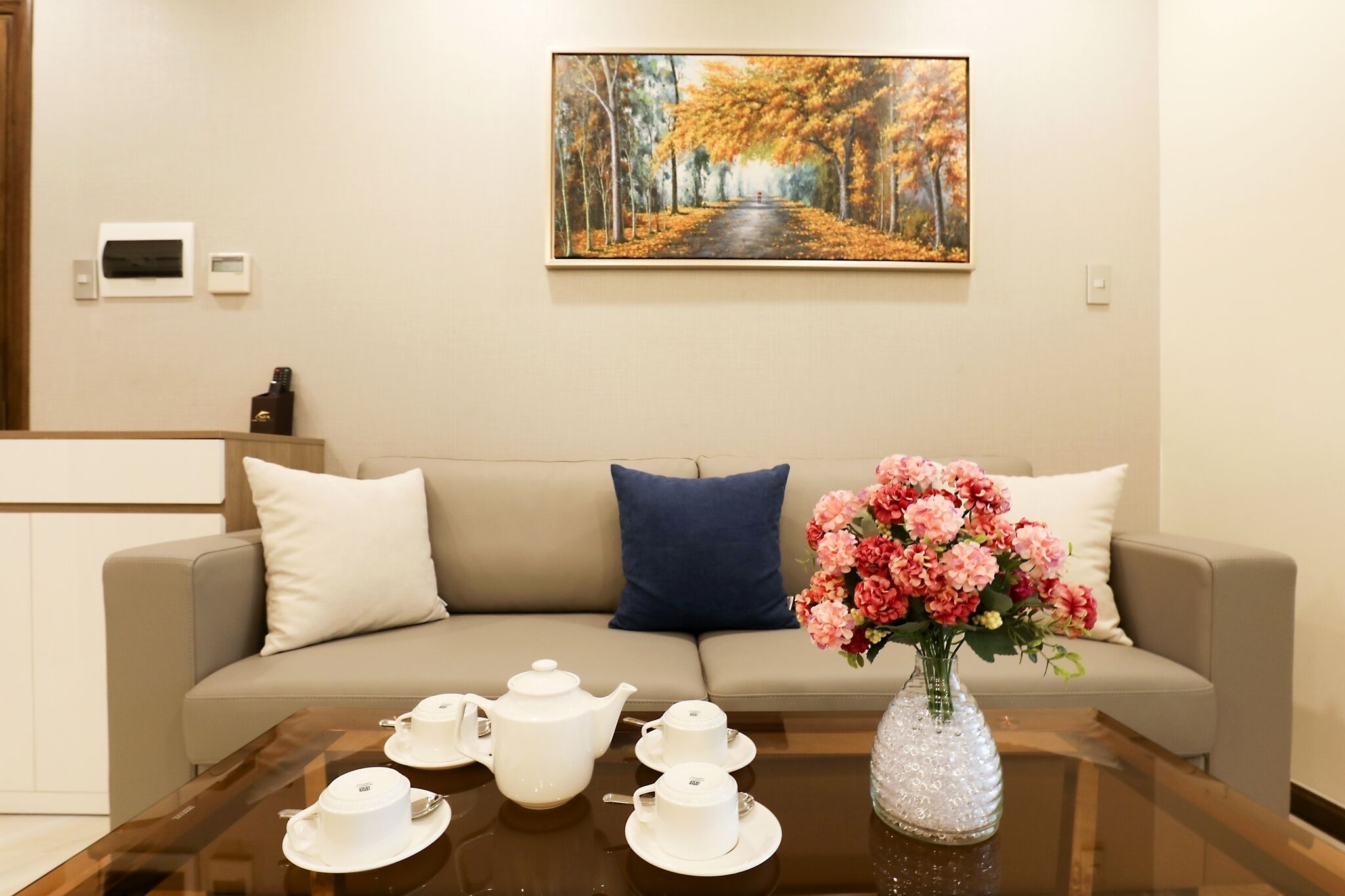 A modern hotel room with a beige sofa, a coffee table with tea sets, a vase of flowers, and a painting of a tree-lined path.