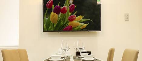 A dining area with a table set for two, a wall-mounted TV, and a floral arrangement on the table.