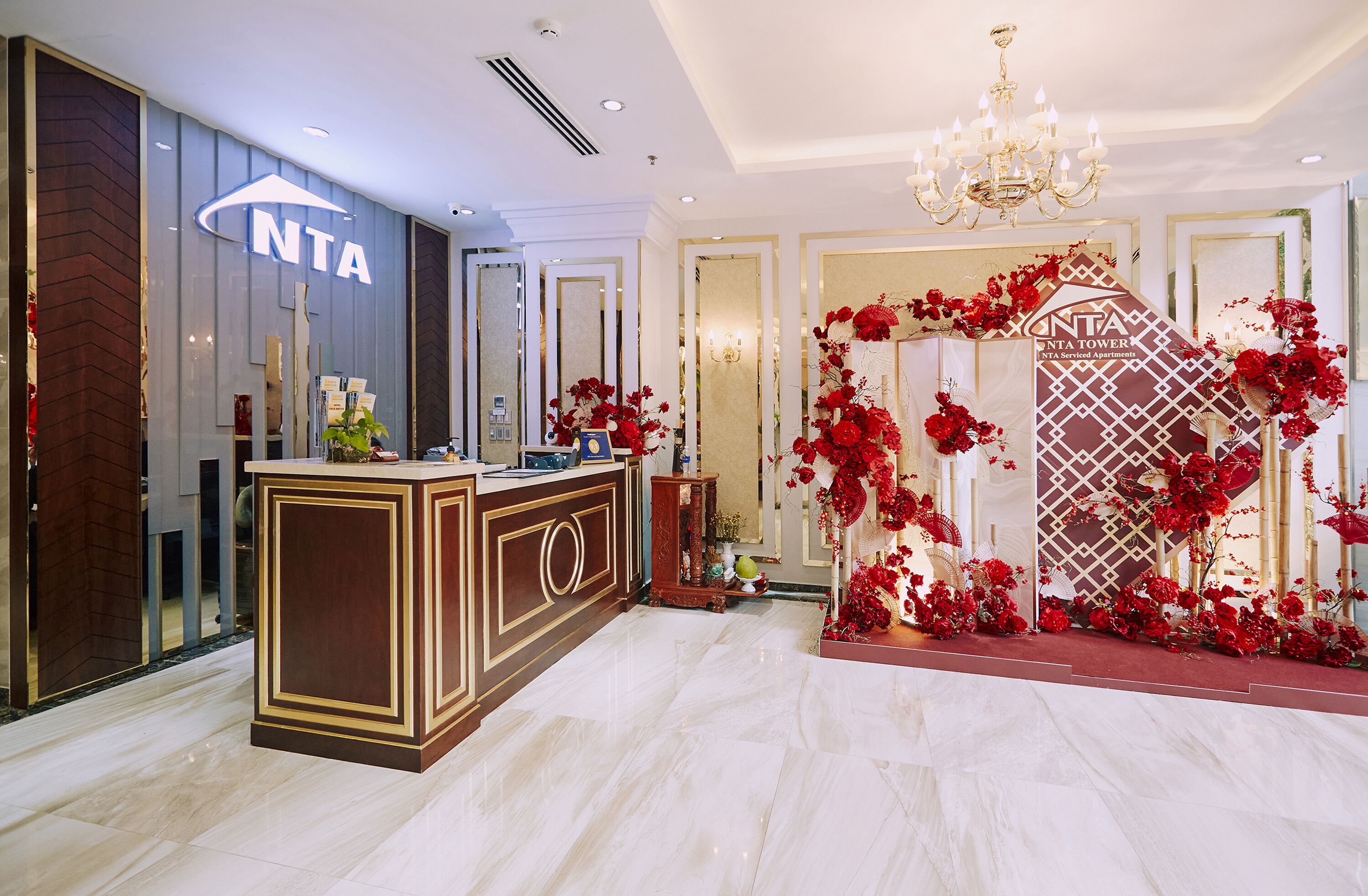 A hotel reception area with a modern design, featuring a reception desk, a chandelier, and red floral decorations.