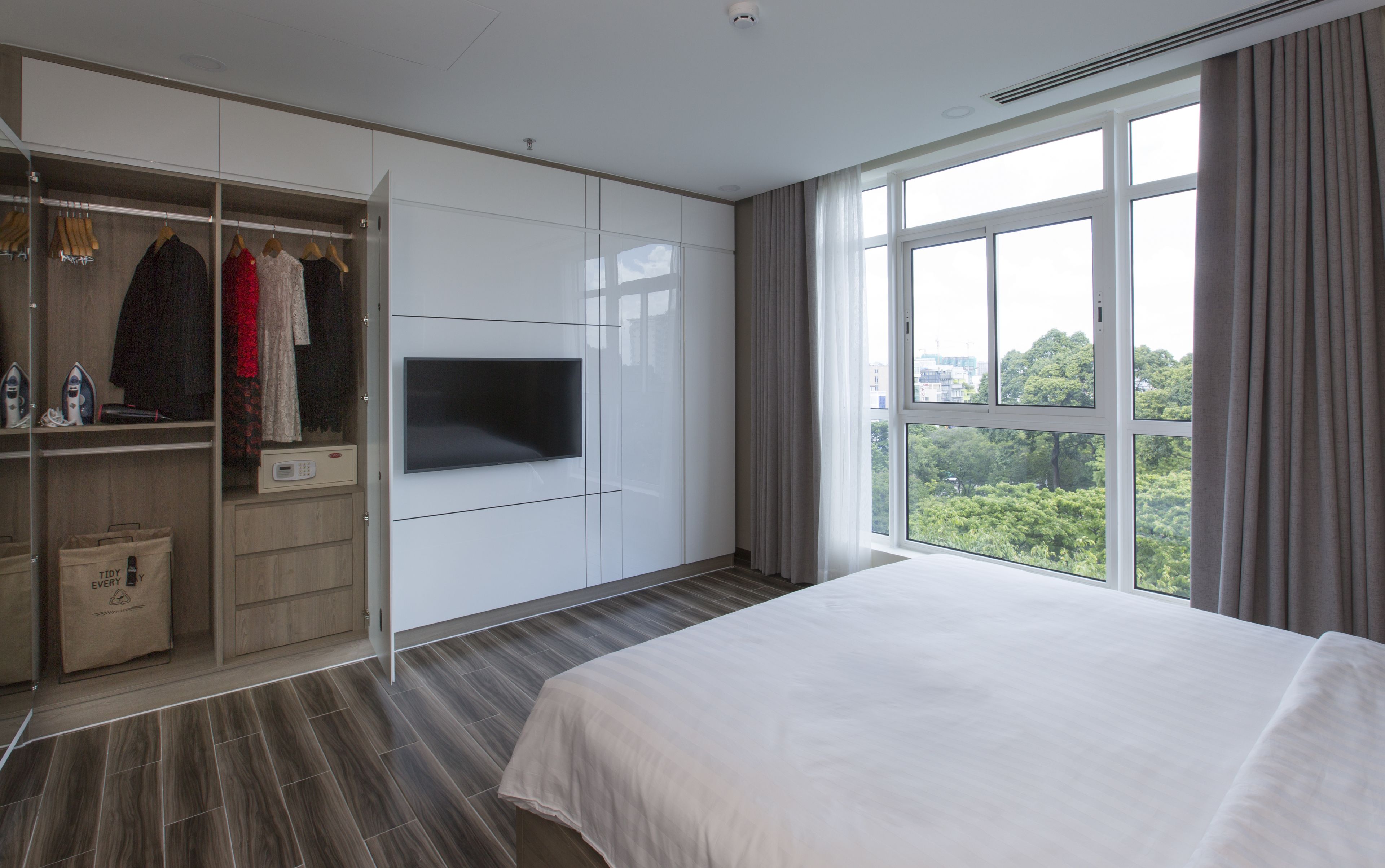 Habitación de hotel moderna con una cama grande, un armario con ropa colgada, un televisor de pantalla plana y vistas a zonas verdes a través de amplios ventanales.