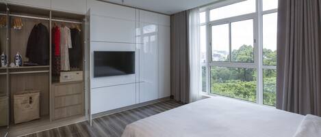 Habitación de hotel moderna con una cama grande, un armario con ropa colgada, un televisor de pantalla plana y vistas a zonas verdes a través de amplios ventanales.