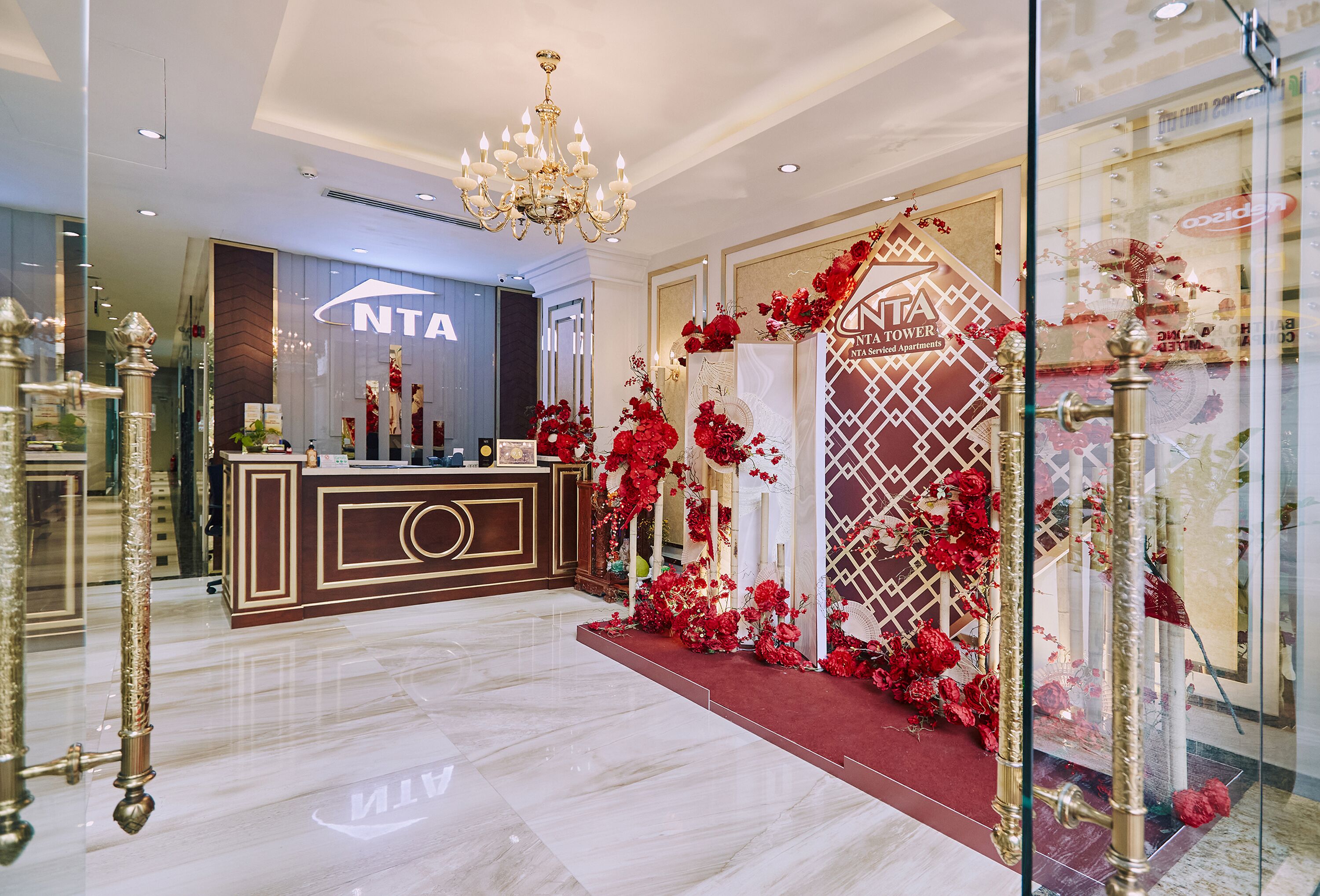 Un vestíbulo de hotel con una recepción, una araña y decoraciones florales rojas.