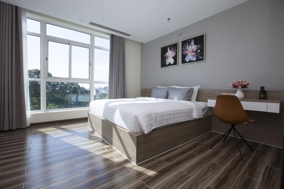 A modern bedroom with a large bed, a desk, and a chair. There are two paintings on the wall and a window with curtains.