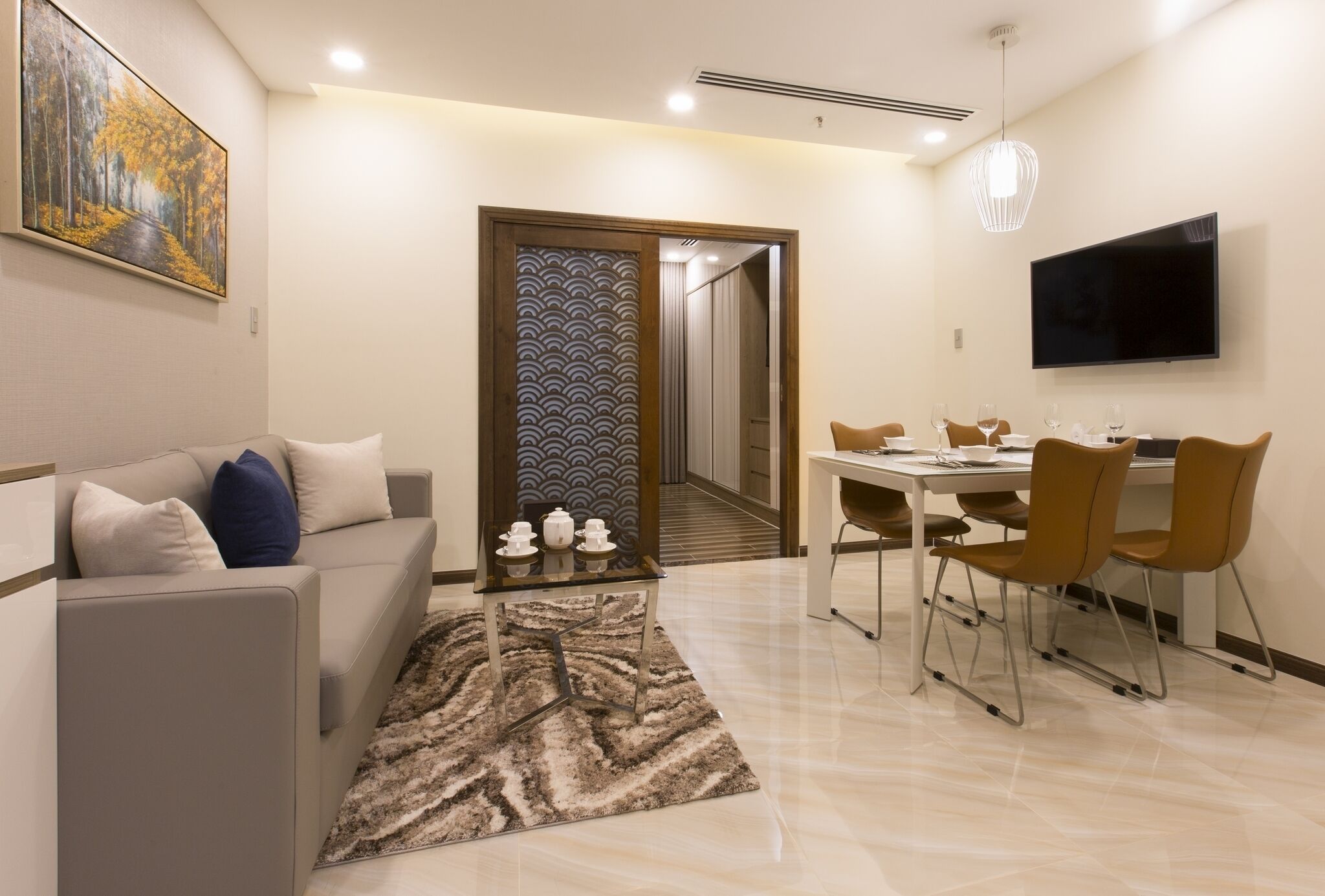 A modern living room with a sofa, a dining table with chairs, a TV mounted on the wall, and a patterned rug.
