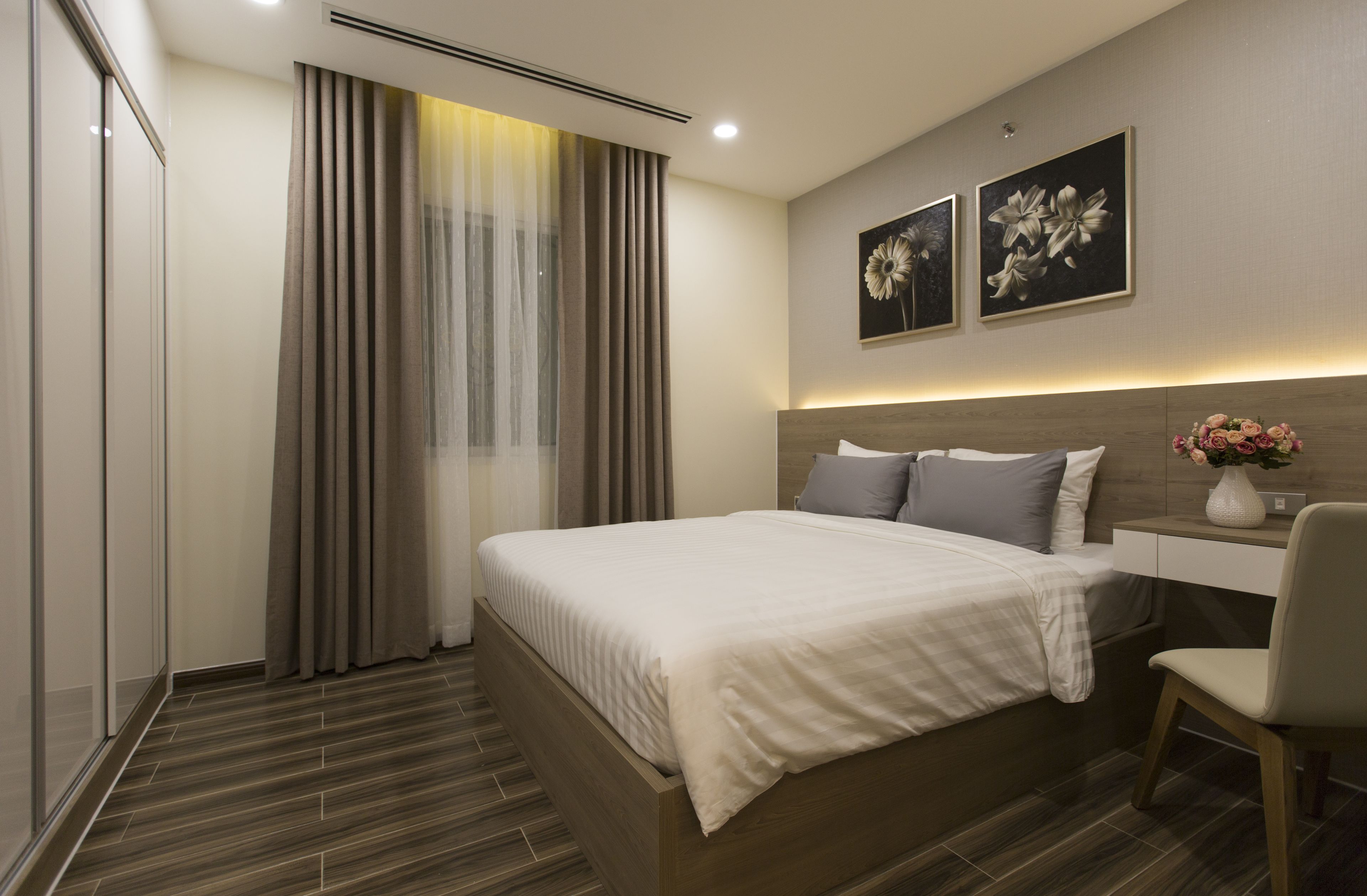 A modern hotel room with a large bed, a bedside table with a vase of flowers, and two framed floral artworks on the wall.