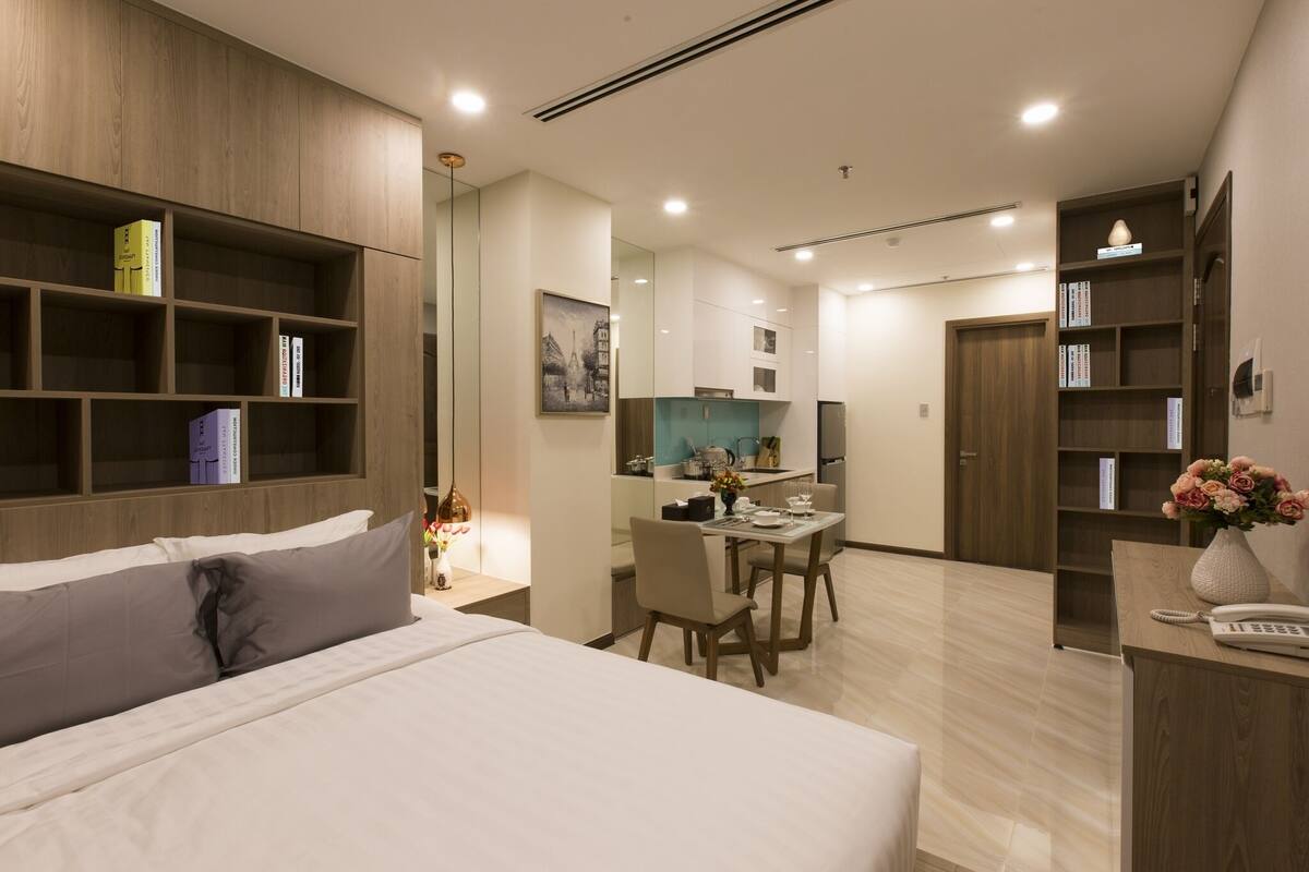 A modern hotel room with a large bed, a desk with chairs, a kitchenette, and a bookshelf.