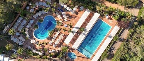 An aerial view of a resort with a large rectangular pool, a smaller circular pool, and multiple sun loungers and umbrellas.