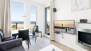 Ein modernes Hotelzimmer mit großem Fenster und Blick auf Strand und Sonnenuntergang. Es gibt einen Essbereich mit Tisch und Stühlen, einen flachbildfernseher an der Wand und einen Kamin.