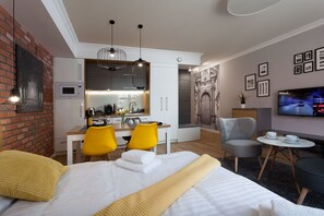 Una habitación de hotel moderna con una cama, un escritorio, dos sillas amarillas, un sillón gris, una mesita redonda y un televisor en la pared.