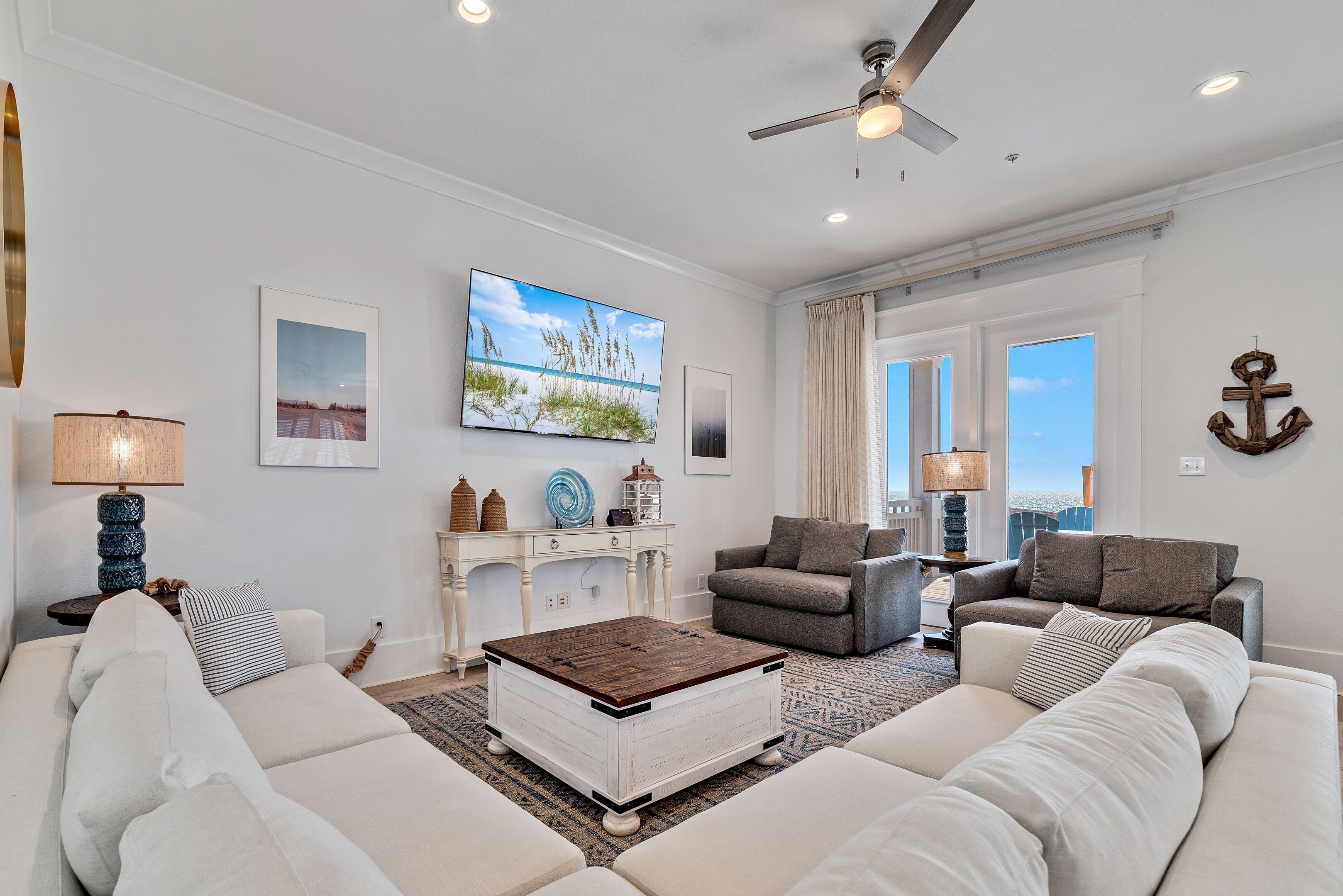 A spacious living room with a large flat-screen TV, a ceiling fan, and a view of the city through large windows.