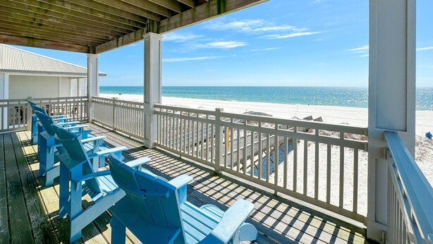 Een overdekt terras met blauwe Adirondack-stoelen, een witte reling en uitzicht op het strand en de oceaan.