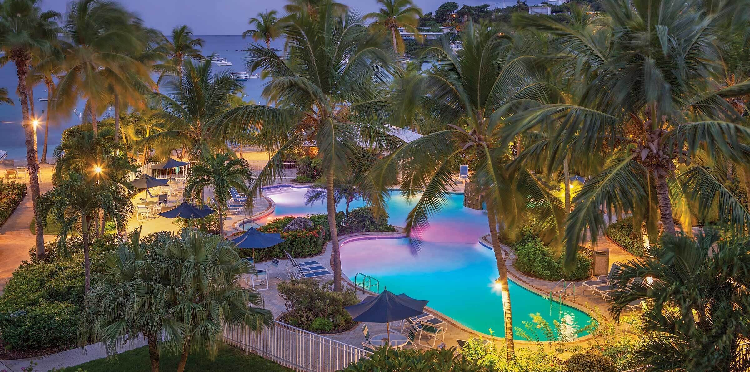 A resort pool area with palm trees, lounge chairs, and a view of the ocean at dusk.