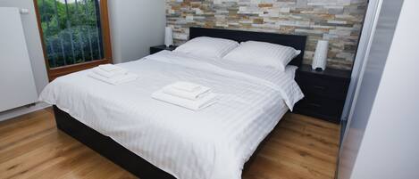 A modern bedroom with a wooden floor, a large bed with white linens, a stone-patterned headboard, and a nightstand with a lamp.