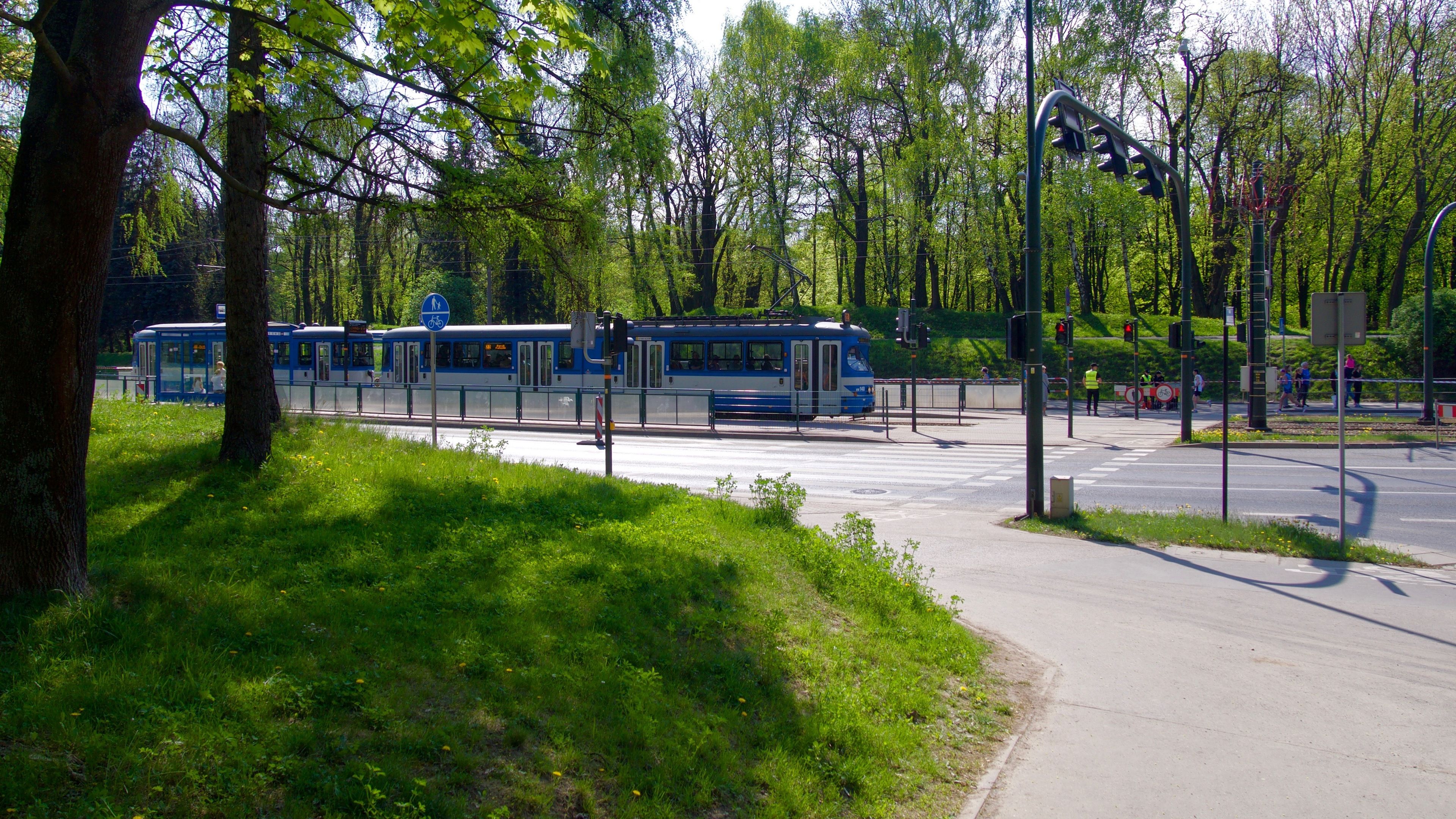 Une station de tram avec un tram bleu, un passage piéton et une zone herbeuse avec des arbres.