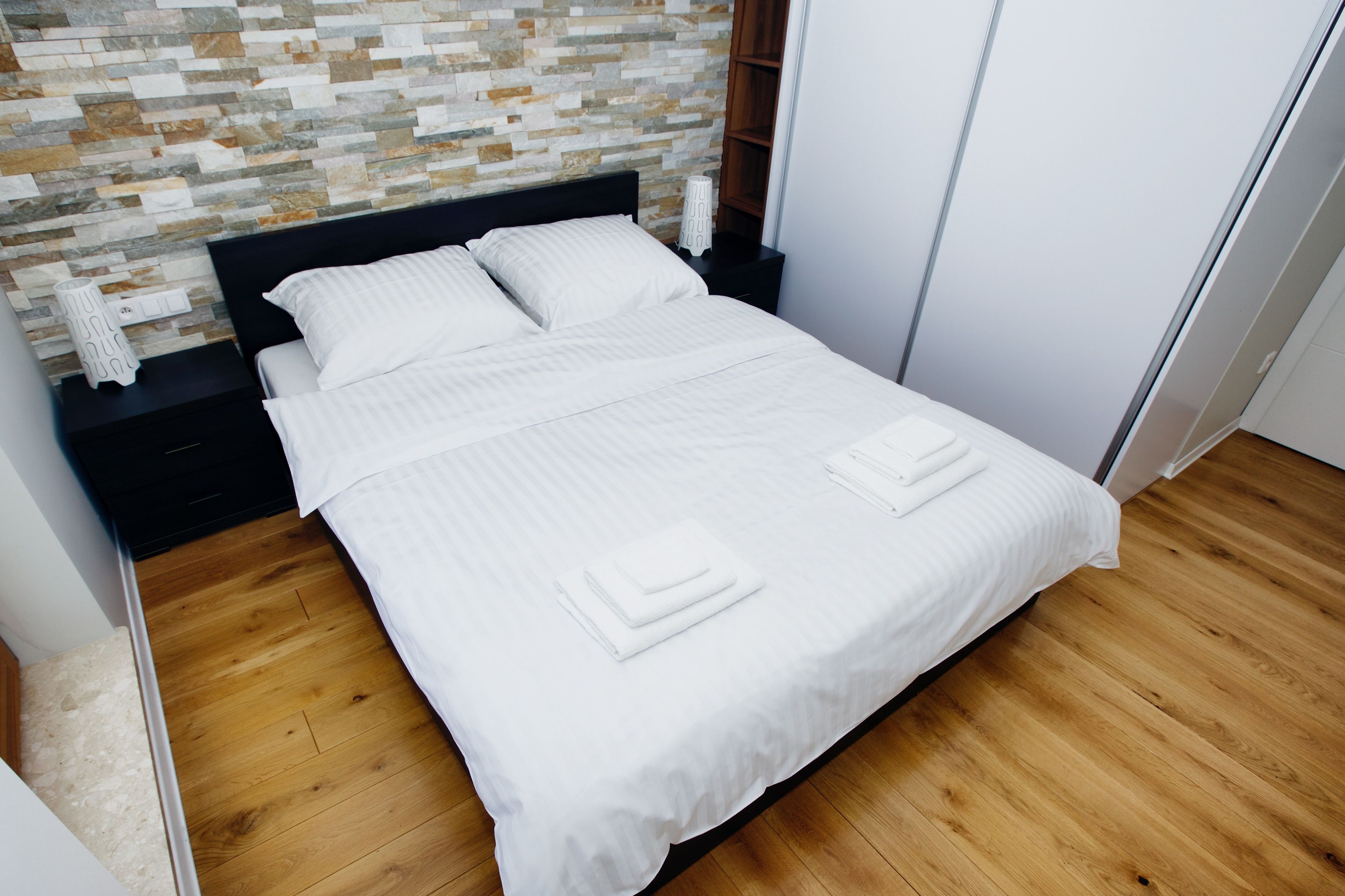Uma cama bem arrumada com lençóis e toalhas brancas, piso de madeira e parede com padrão de pedra.