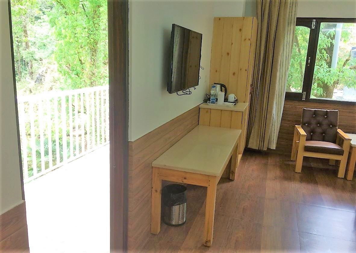A room with a wooden bench, a television mounted on the wall, a small table with a kettle, and a chair.