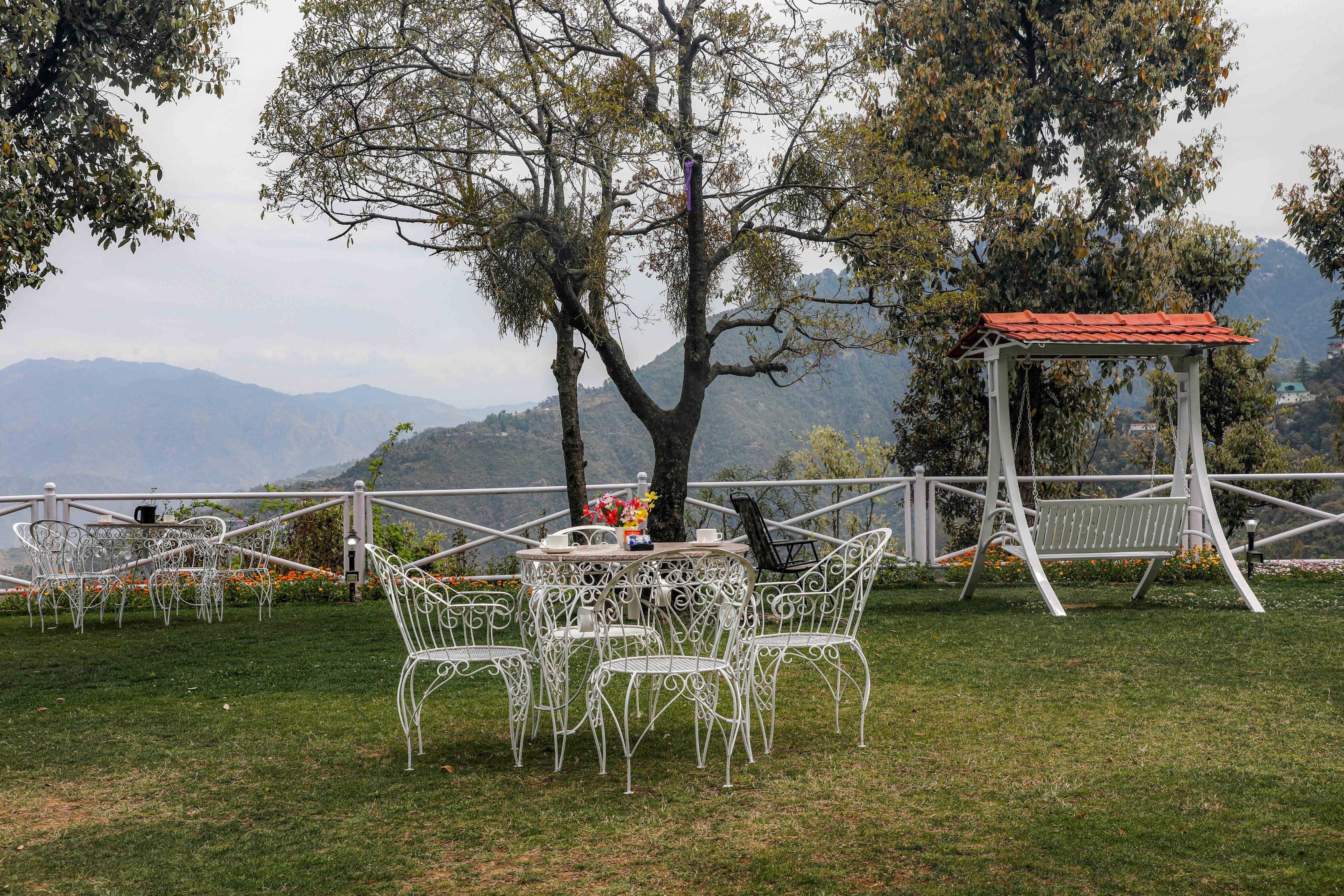 An outdoor seating area with white metal chairs and tables, surrounded by a white fence, overlooking a mountainous landscape.