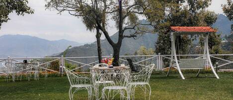An outdoor seating area with white metal chairs and tables, surrounded by a white fence, overlooking a mountainous landscape.