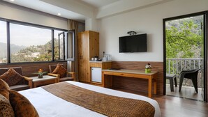 A hotel room with a large bed, a desk, a TV, and a balcony with a view of the hills. - The Mountain Quail (Dehradun)