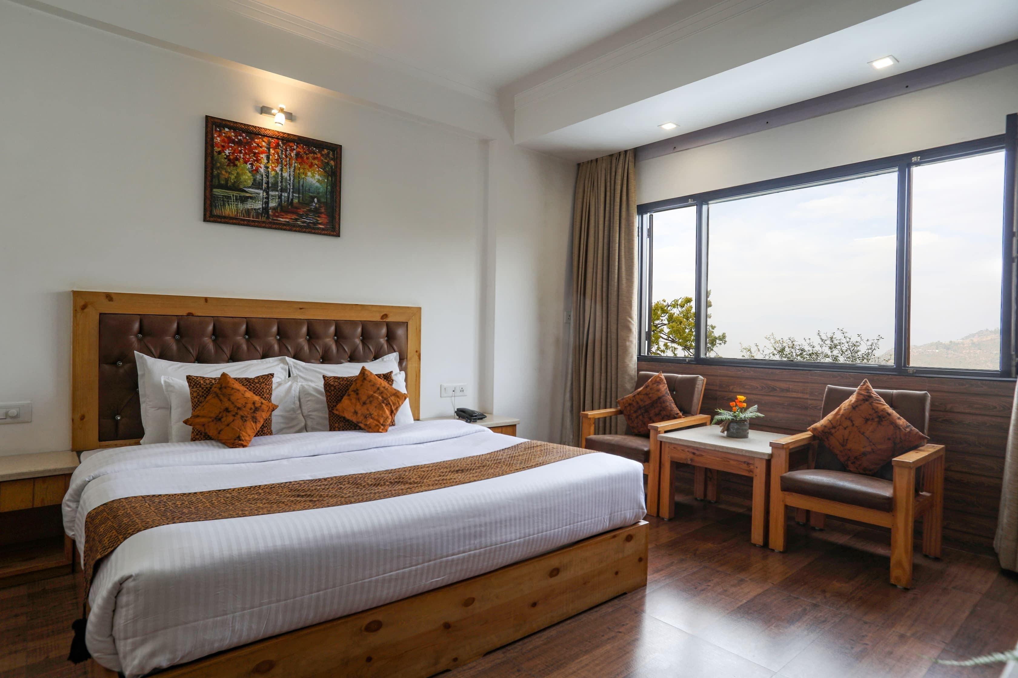 Habitación de hotel con una cama grande, un cabecero de madera, una mesita pequeña, dos sillas y un ventanal con vistas.