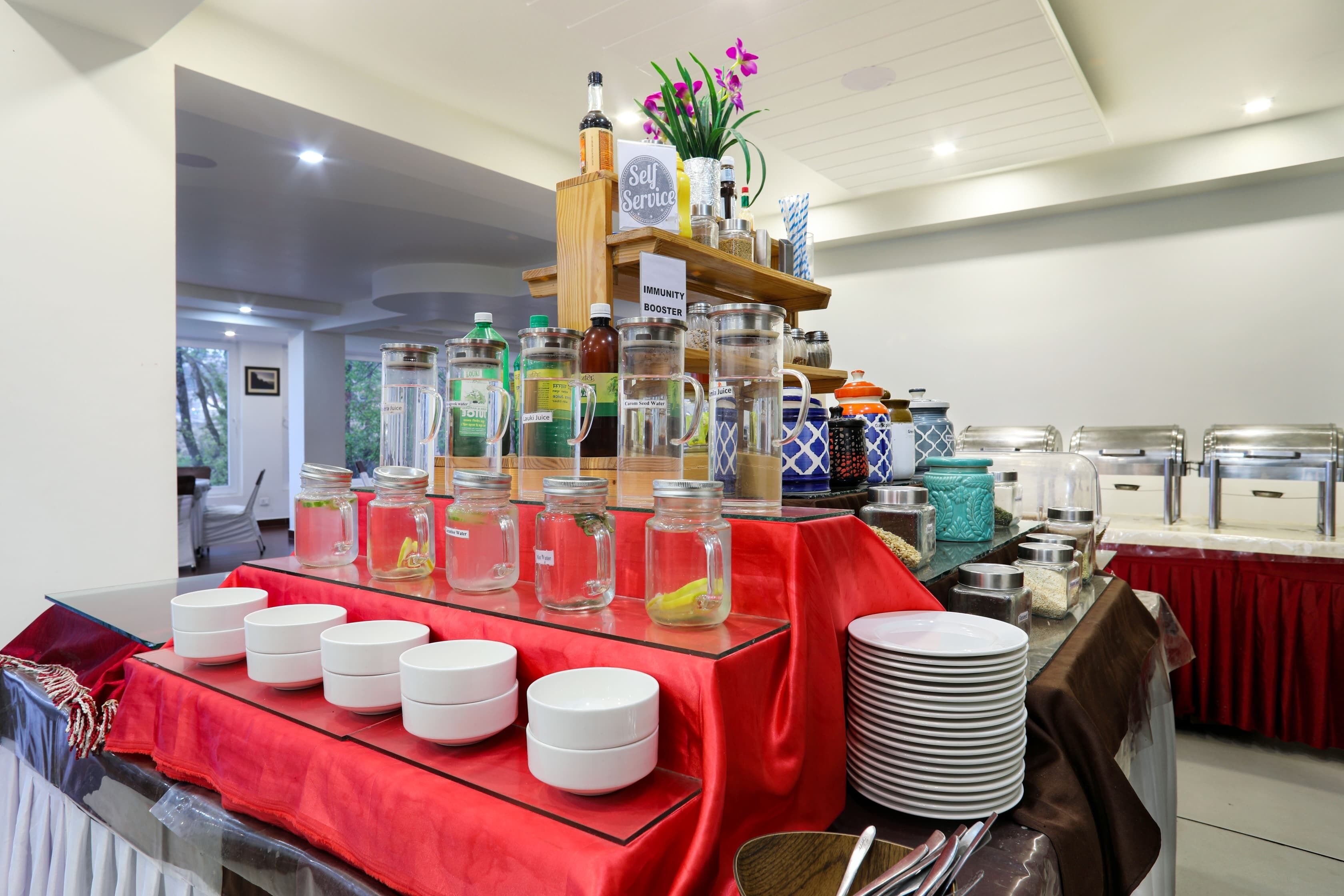 Tarjolla on erilaisia ruokia ja juomia sisältäviä astioita, ja ne on järjestetty buffet-tyylisesti. Lisäksi paikalla on kyltti, jossa kerrotaan, että ruoat ja juomat ovat asiakkaiden itsenäisesti valittavissa, sekä koristeellinen hylly, jossa on kukkia.