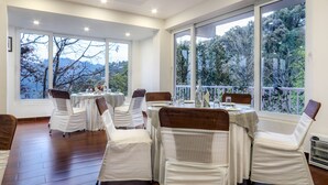 A dining area with tables set for six, large windows offering a view of trees and mountains, and a wooden floor. - The Mountain Quail (Dehradun)
