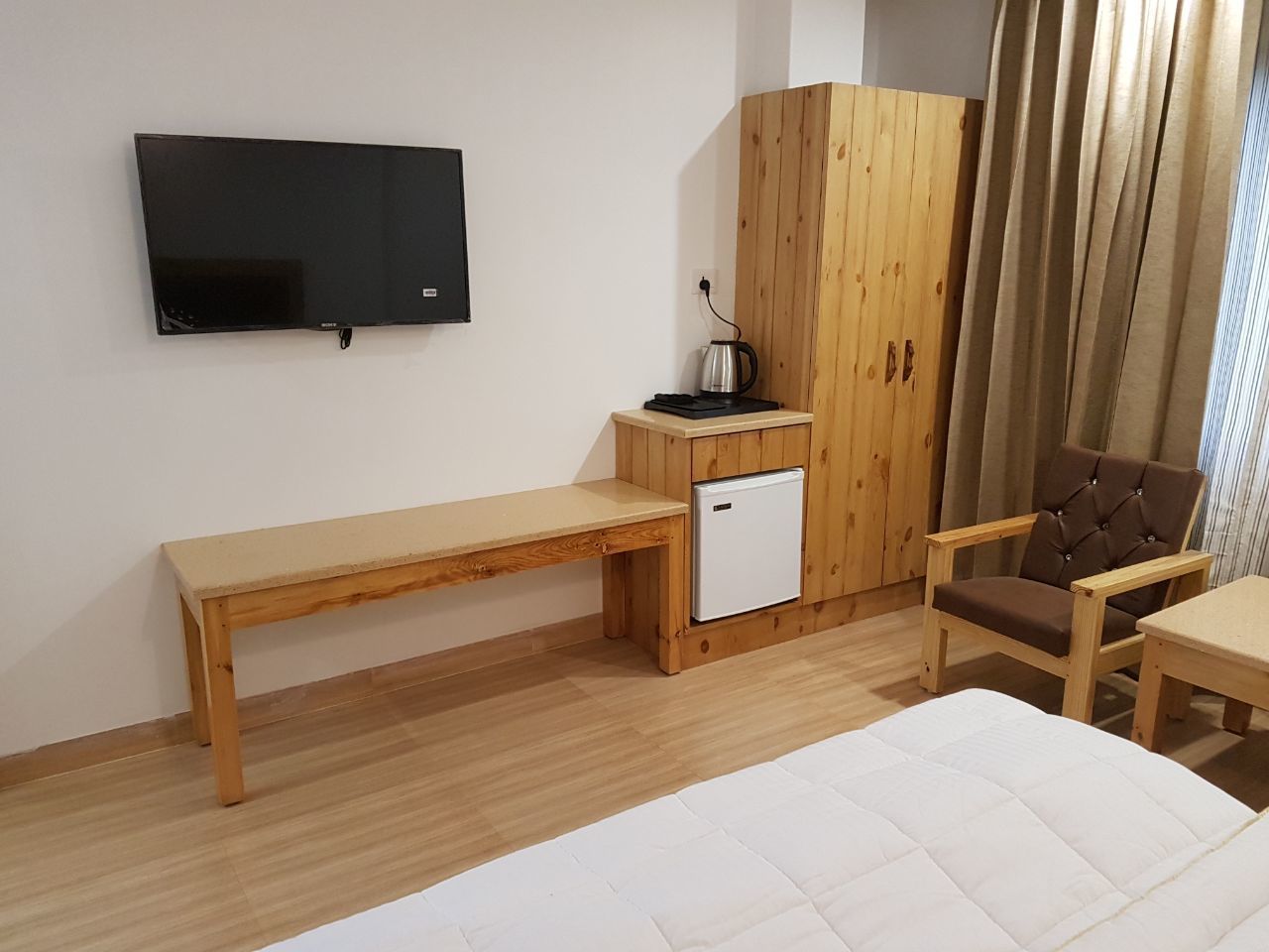 A room with a bed, a wooden bench, a small white fridge, a television mounted on the wall, and a wooden cabinet.