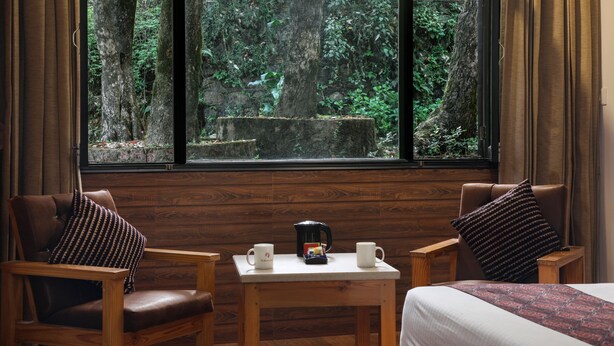 A room with a large window offering a forest view, a bed, two chairs, and a small table with a coffee pot and two mugs. - The Mountain Quail (Dehradun)
