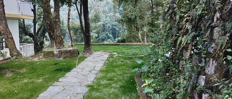 A stone pathway leading through a lush garden with trees and a white building on the left.