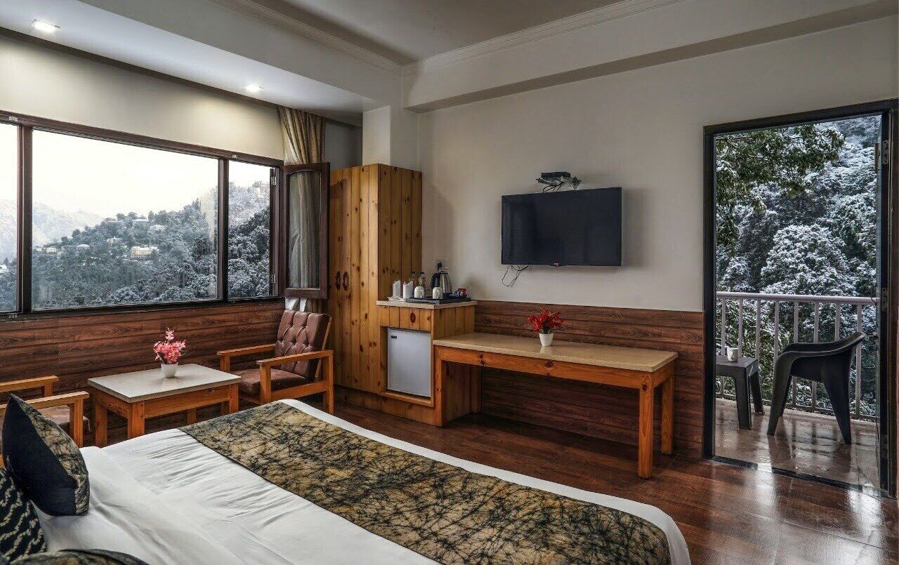 A room with a bed, a wooden desk, a television, and a balcony with a view of snow-covered trees.