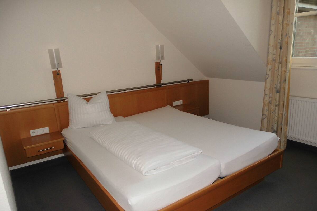 A neatly made bed in a room with a sloped ceiling, a wooden headboard, and a bedside table with a lamp.