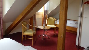 A room with a sloped ceiling, wooden beams, a bed, two chairs, a small table, and a bed with a white mattress. - Landgasthaus Hotel Eggert (Rheine)