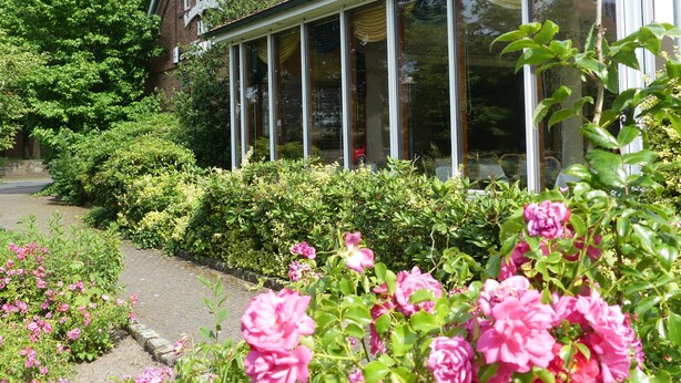 A traditional house with a conservatory, surrounded by lush greenery and flowering plants. - Landgasthaus Hotel Eggert (Rheine)