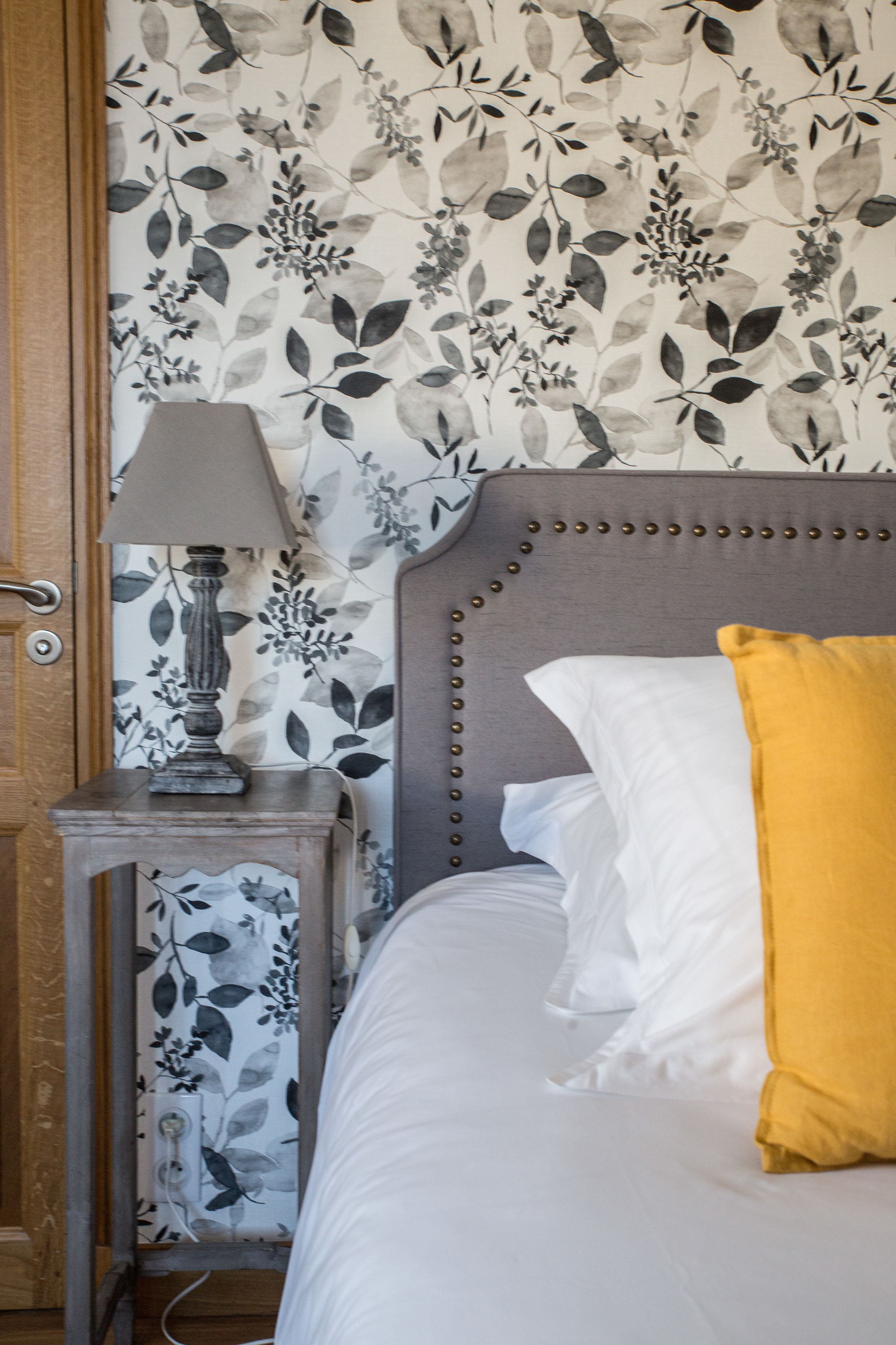 A bedroom with a floral wallpaper, a bedside table with a lamp, and a bed with white and yellow pillows.